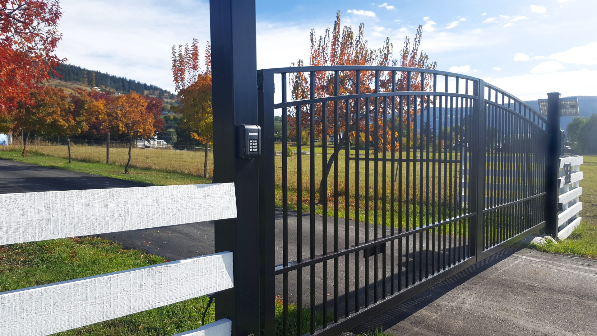 White Wood Fence with Black Iron security gate. Locked with combination. Rural non Urban setting. Red Autumn trees in background.